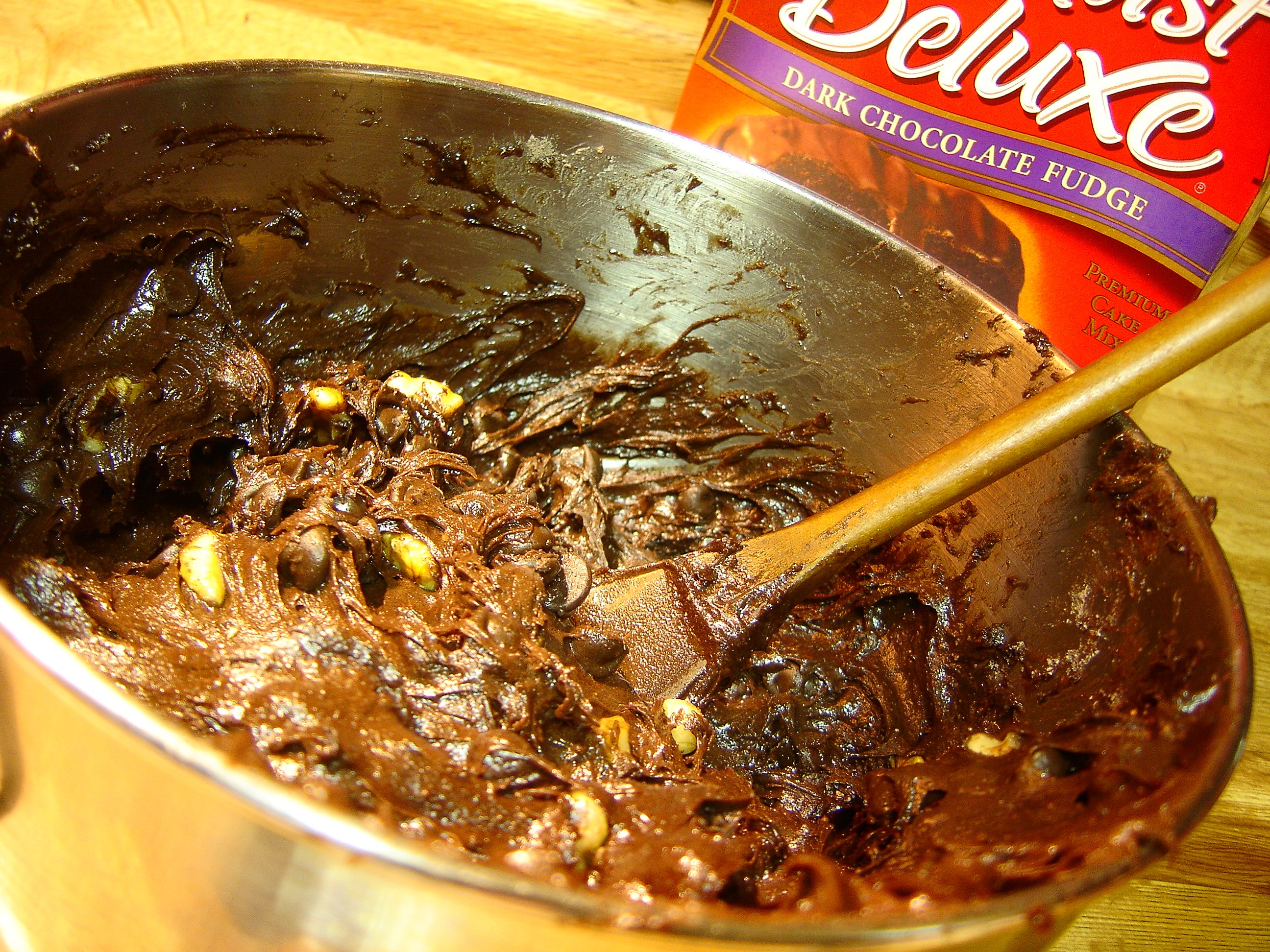 Photograph of a bowl with chocolate cake batter with chocolate chips and nuts in it, with a box of cake mix visible in the background.