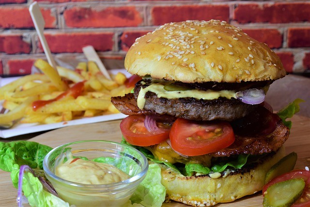 A burger with bacon, tomatoes, lettuce, cheese, onions, and pickles with a side of French fries and mayonnaise for dipping.
