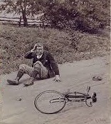A bike lies in the middle of the street. A man sits right next to it, rubbing his head.