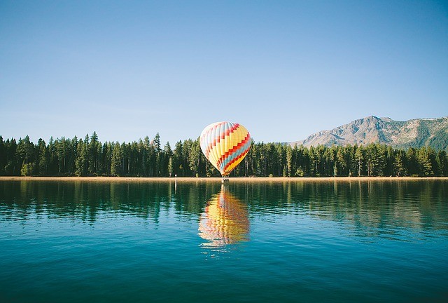 A multicolored hot air balloon resting at the edge of a lake in the view of mountains and a forest