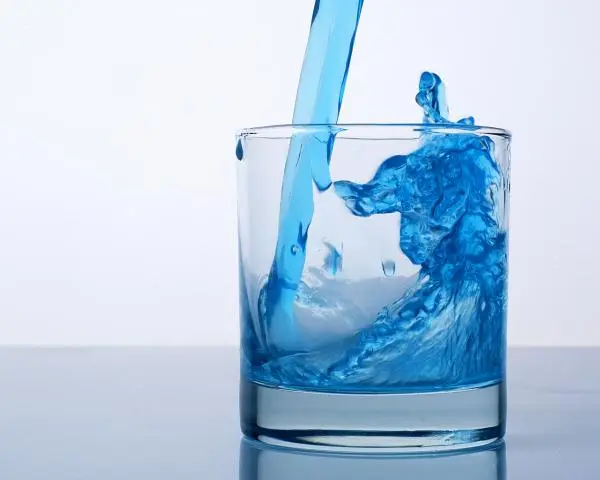 Blue water pours from above into a clear glass and splashes up inside as the glass fills, against a light background.