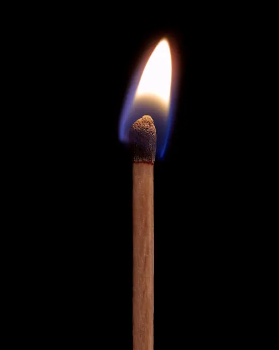 A lit wooden match against a black background: a yellow-white flame with a blue base rises from the burnt match head.