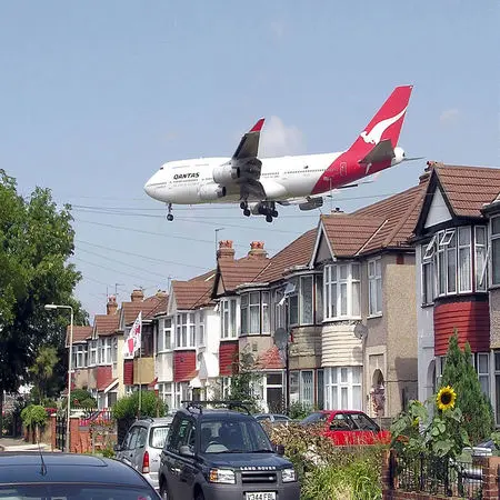 The image shows a large airplane flying directly over a housing plan.